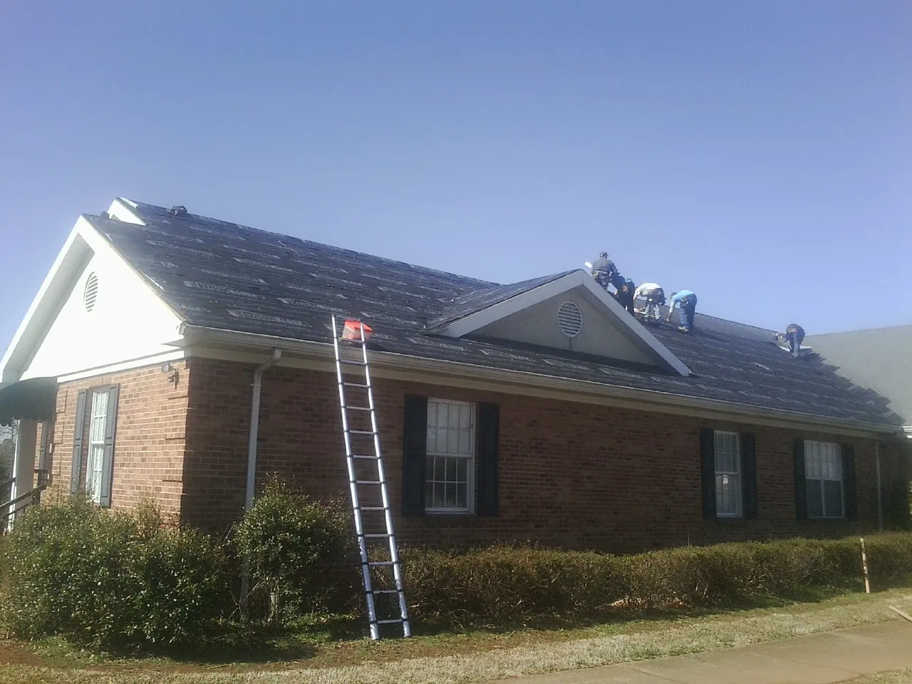 Crew tearing shingles off a brick ranch roof with a ladder set — Metal Roof Repair work by our crew in Everest