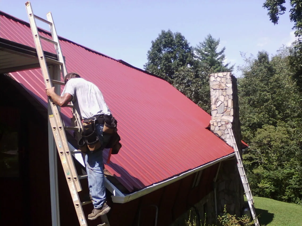 Roofer on a ladder beside a red metal roof and stone chimney — Soffit & Fascia Repair by Roofing Brothers in Everest